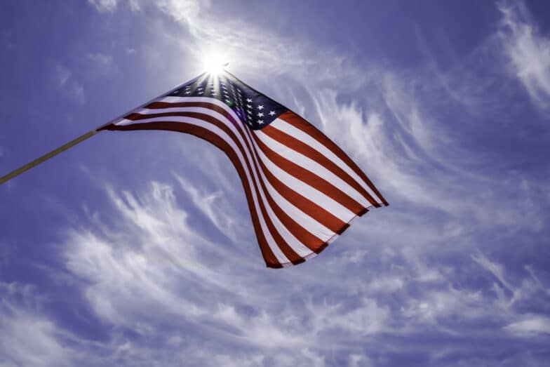 American flag waving in the sky with streaked clouds in a vibrant beautiful blue sky and a sun starburst behind the USA flag. The national red, white and blue star stiped US flag flying high.